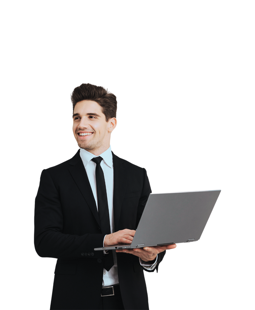 A man wearing a black suit and tie, is smiling while holding and using a laptop.